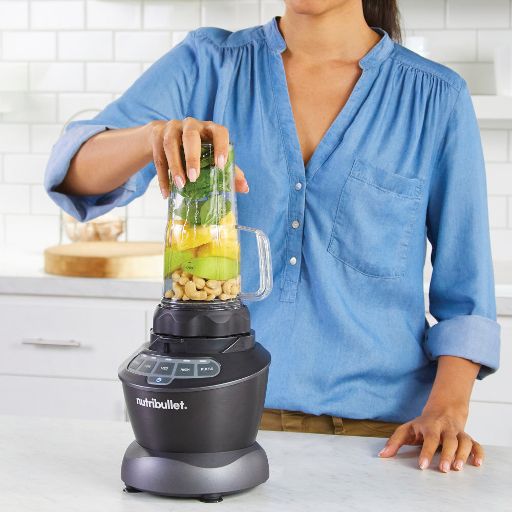Osoba obsługuje blender kielichowy z logo \'nutribullet\', w którym znajdują się owoce i orzechy. W tle widoczna jest biała kuchnia z kafelkami na ścianie.