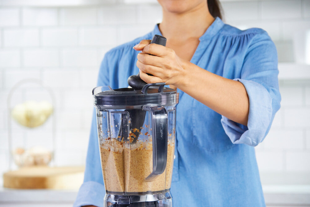 Osoba w niebieskiej koszuli obsługuje blender kielichowy z ciemnym uchwytem, w którym znajduje się brązowa mieszanka. W tle widoczna jest jasna kuchnia z białymi kafelkami.
