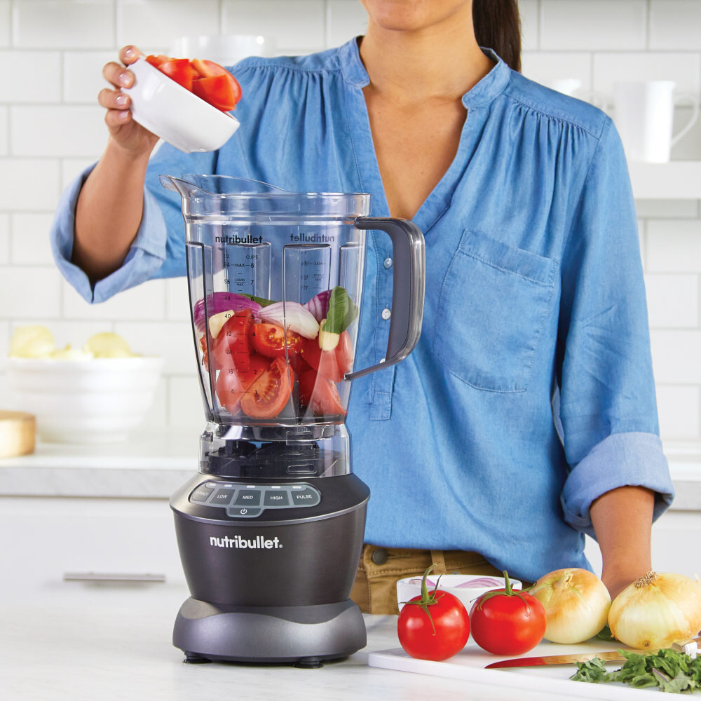Osoba w niebieskiej koszuli dodaje pokrojone pomidory do blendera kielichowego marki nutribullet, w którym znajdują się już inne warzywa. Na blacie obok leżą pomidory i cebule.