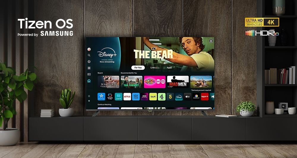 Telewizor wyświetla ekran główny z aplikacjami takimi jak Disney+, Netflix i YouTube. Na ścianie obok telewizora widoczne jest logo \'Tizen OS Powered by Samsung\'.