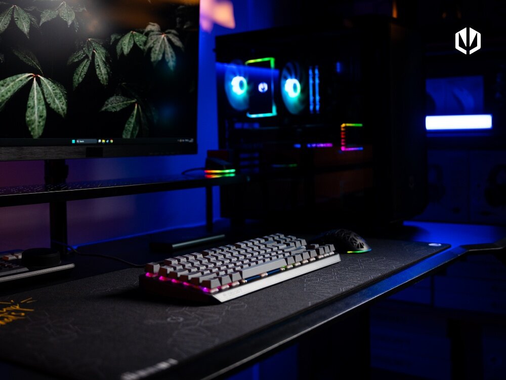Na biurku znajduje się klawiatura mechaniczna z podświetleniem RGB oraz mysz komputerowa. W tle widać monitor z wyświetlonymi liśćmi oraz komputer z podświetlonymi wentylatorami.