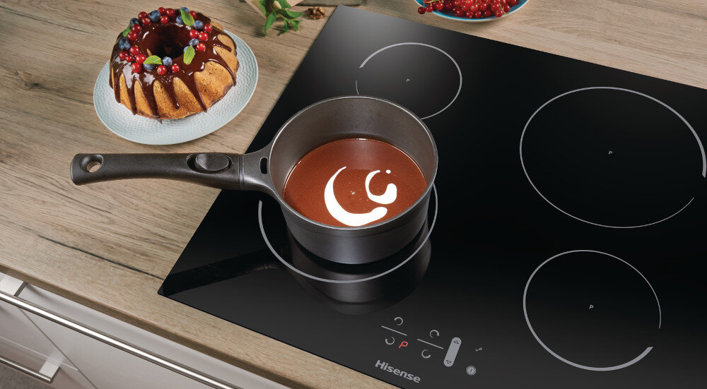 Czarna płyta indukcyjna z czterema polami grzewczymi, na której stoi rondel z czekoladą. Obok na blacie znajduje się ciasto z owocami, a na płycie widoczne jest logo \'Hisense\'.