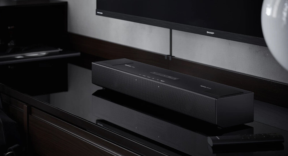 Na obrazie znajduje się czarny soundbar umieszczony na ciemnym, błyszczącym blacie, przed telewizorem z logo SHARP. Obok soundbara leży pilot.