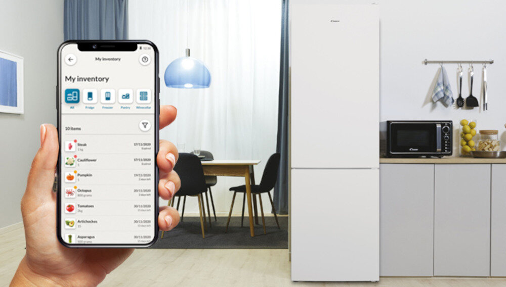 Osoba trzyma smartfon z aplikacją pokazującą listę produktów spożywczych. W tle znajduje się biała lodówka, obok której stoi mikrofalówka i kilka kuchennych akcesoriów.