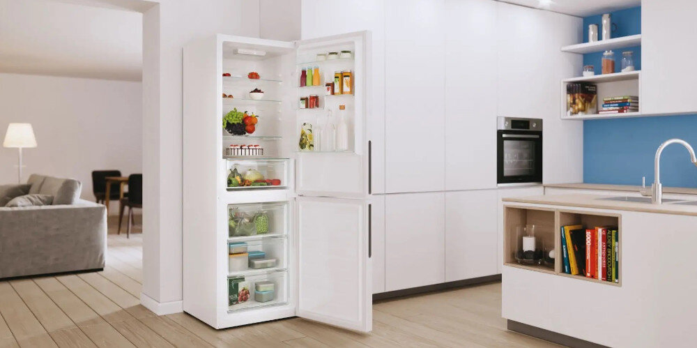Wnętrze nowoczesnej kuchni z otwartą białą lodówką wypełnioną różnorodnymi produktami spożywczymi. Obok lodówki znajduje się zabudowa kuchenna z piekarnikiem i zlewem.