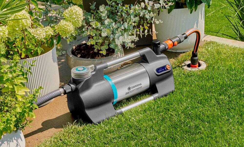 Na trawniku znajduje się elektryczna pompa ogrodowa GARDENA Silent Comfort 09059-20 6500, podłączona do węża ogrodowego. W tle widoczne są donice z roślinami.