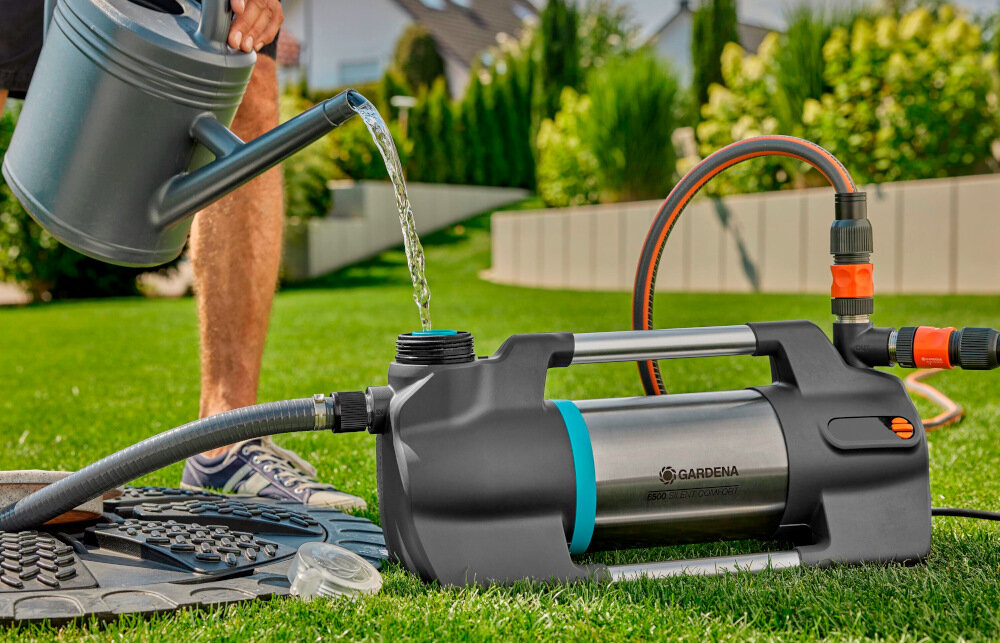 Na trawniku znajduje się elektryczna pompa ogrodowa GARDENA Silent Comfort 09059-20 6500 z podłączonymi wężami. Obok osoba nalewa wodę z konewki do otworu pompy.