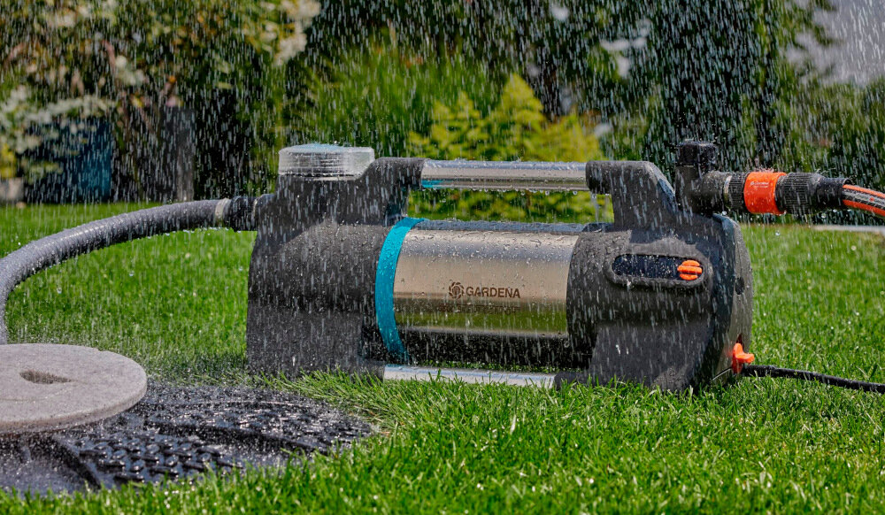 Na obrazie widoczna jest pompa ogrodowa z napisem \'GARDENA\', umieszczona na trawie, z podłączonym wężem ogrodowym. W tle widać roślinność, a na pompę pada woda.