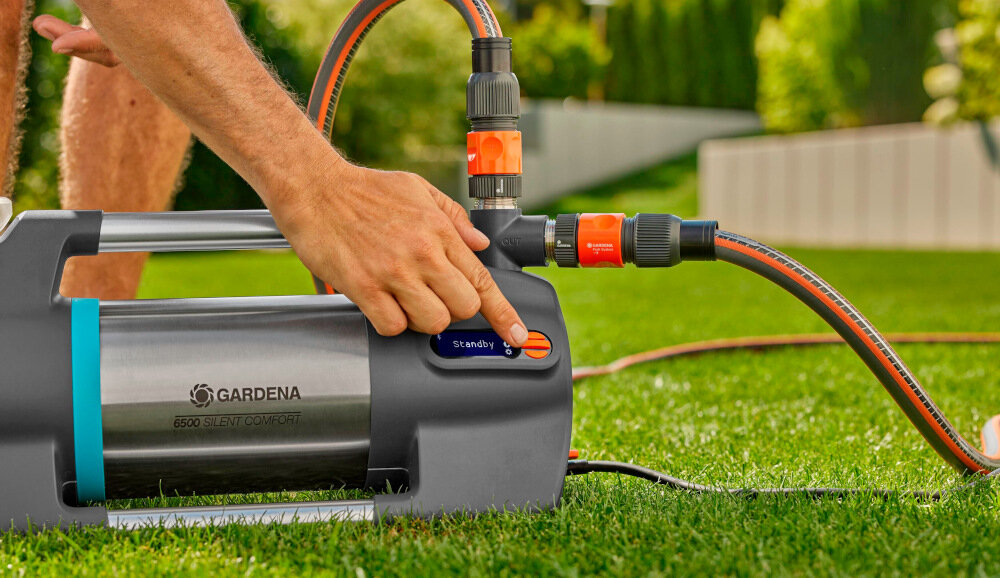 Osoba obsługuje elektryczną pompę ogrodową GARDENA Silent Comfort 6500, naciskając przycisk oznaczony jako \'stand-by\'. Pompa jest podłączona do węża ogrodowego i znajduje się na trawie.