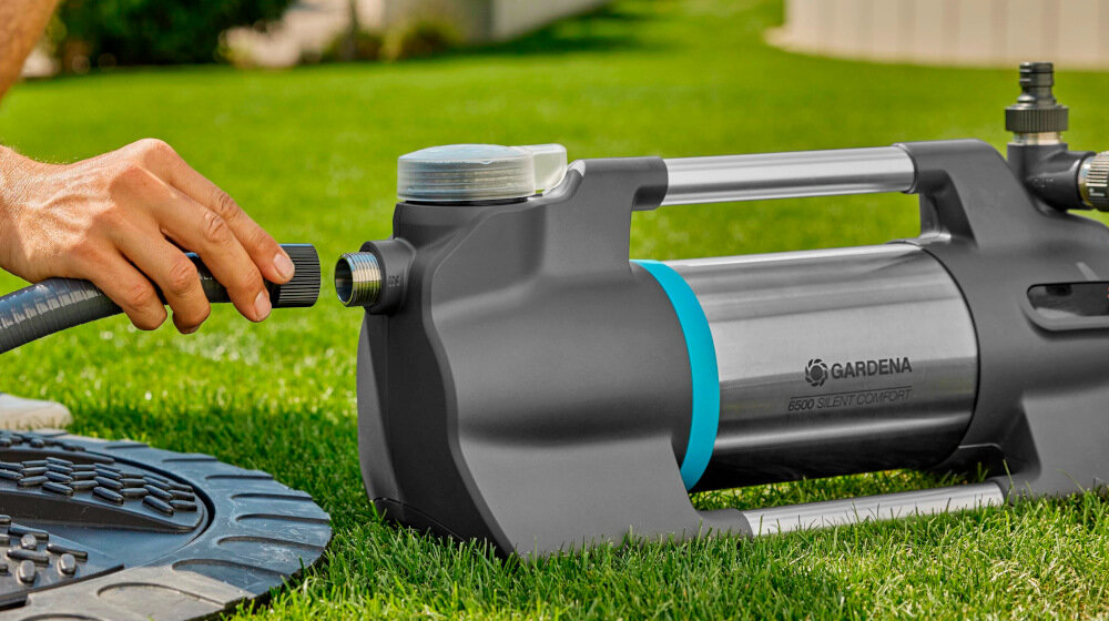 Osoba podłącza wąż do elektrycznej pompy ogrodowej GARDENA Silent Comfort 6500, która znajduje się na trawie. Na pompie widoczne jest logo GARDENA.