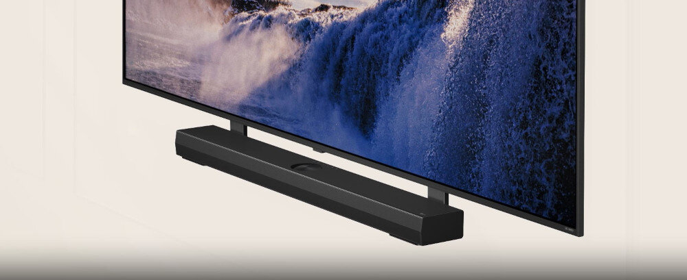 Na obrazie widoczny jest czarny soundbar umieszczony pod telewizorem, na którym wyświetlany jest obraz fal morskich.