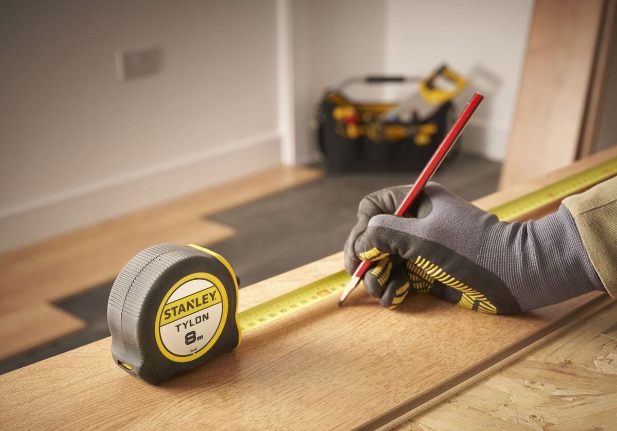 Miara zwijana STANLEY 0-30-657 (8 m), precyzja, taśma, zwijanie, blokada, Ręka w szarej rękawicy trzyma zwiniętą miarę Stanley Tylon 8m, obok czerwony ołówek na drewnianej podłodze i skrzynka z narzędziami w tle.