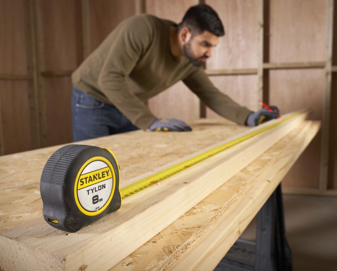 Miara zwijana STANLEY 0-30-657 (8 m), trwała, taśma, Tylon, odporność, Mężczyzna w roboczym ubraniu i rękawicach mierzy deskę drewnianą żółtą miarą Stanley Tylon 8m x 25mm na stole warsztatowym, obok leży zwinięta czarna obudowa z żółtym logo.