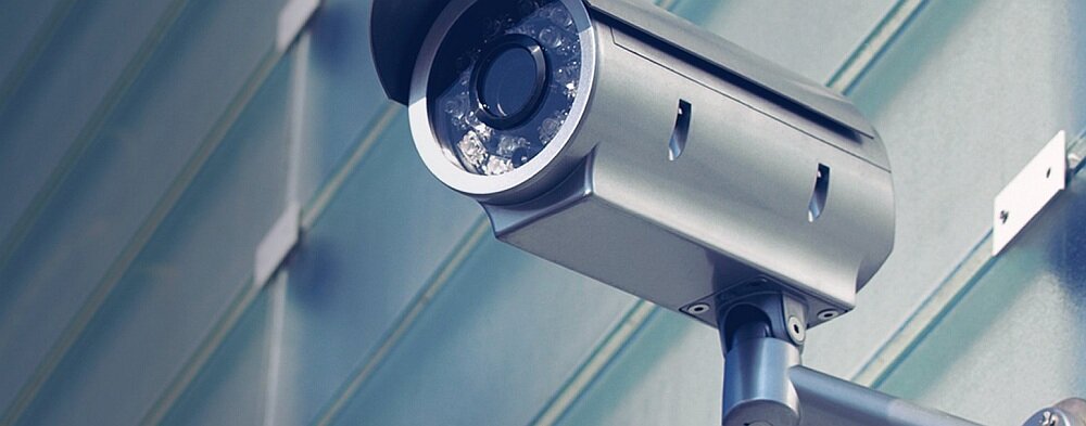Obraz przedstawia metalową kamerę monitoringu zamontowaną na ścianie zewnętrznej budynku. Kamera jest skierowana w dół i ma cylindryczny kształt.