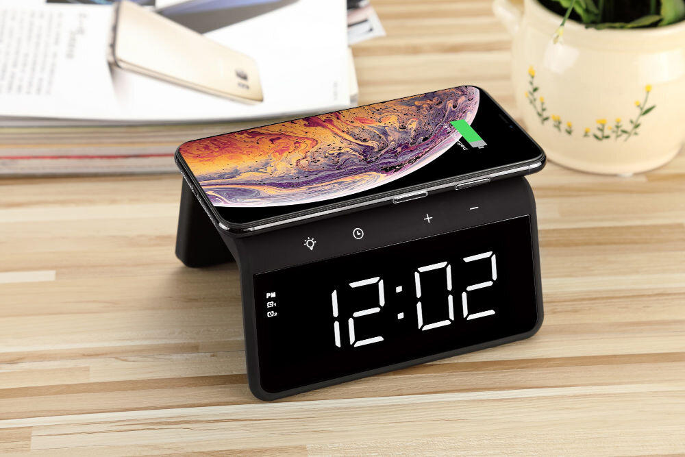 Na drewnianym blacie stoi czarny budzik z wyświetlaczem pokazującym godzinę 12:02, na którym leży smartfon z widocznym wskaźnikiem ładowania. W tle znajduje się stos czasopism i doniczka z rośliną.