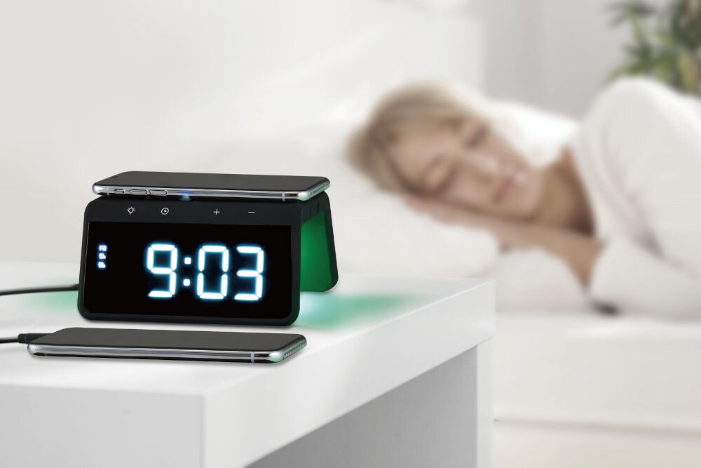 Na białym stoliku nocnym znajduje się czarny budzik z wyświetlaczem pokazującym godzinę 9:03. Obok budzika leży smartfon, a w tle widać osobę leżącą na łóżku.