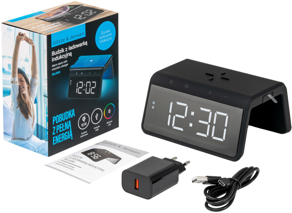 Na obrazie znajduje się czarny budzik z dużym wyświetlaczem cyfrowym pokazującym godzinę 12:30, obok którego leży ładowarka i kabel USB. W tle widoczny jest karton z napisem \'Gotze & Jensen Budzik z ładowarką indukcyjną\' oraz \'Pobudka z pełną energią\'.