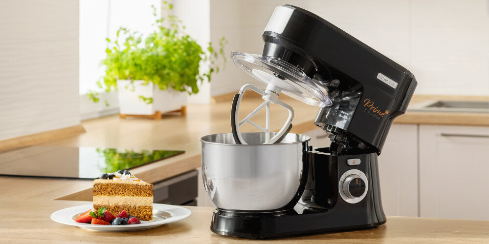Czarny robot kuchenny planetarny z metalową misą stoi na blacie kuchennym. Obok znajduje się kawałek ciasta z owocami na talerzu.