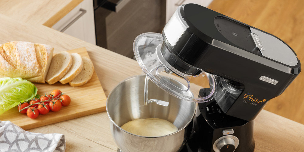 Czarny robot kuchenny z metalową misą, w której znajduje się ciasto. Obok na blacie leży deska z pokrojonym chlebem, pomidorkami i liściem sałaty.