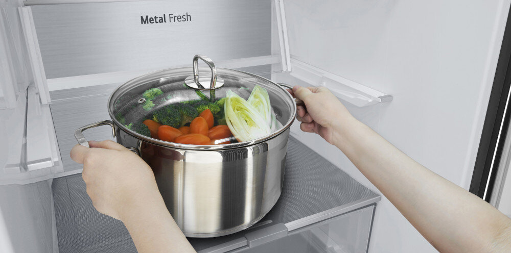 Osoba trzyma garnek z warzywami przed otwartą lodówką z napisem \'Metal Fresh\'.