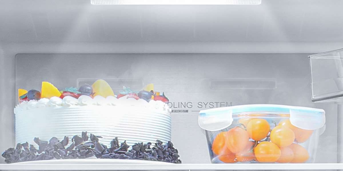 Wnętrze lodówki z widocznym tortem udekorowanym owocami oraz przezroczystym pojemnikiem z pomarańczowymi owocami. Na ścianie lodówki widoczny jest napis \'COOLING SYSTEM\'.