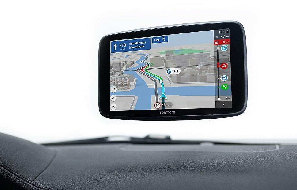 Na zdjęciu widać nawigację samochodową z mapą na ekranie, pokazującą trasę z informacjami o odległości i czasie. Na dole ekranu widnieje logo \'TomTom\'.