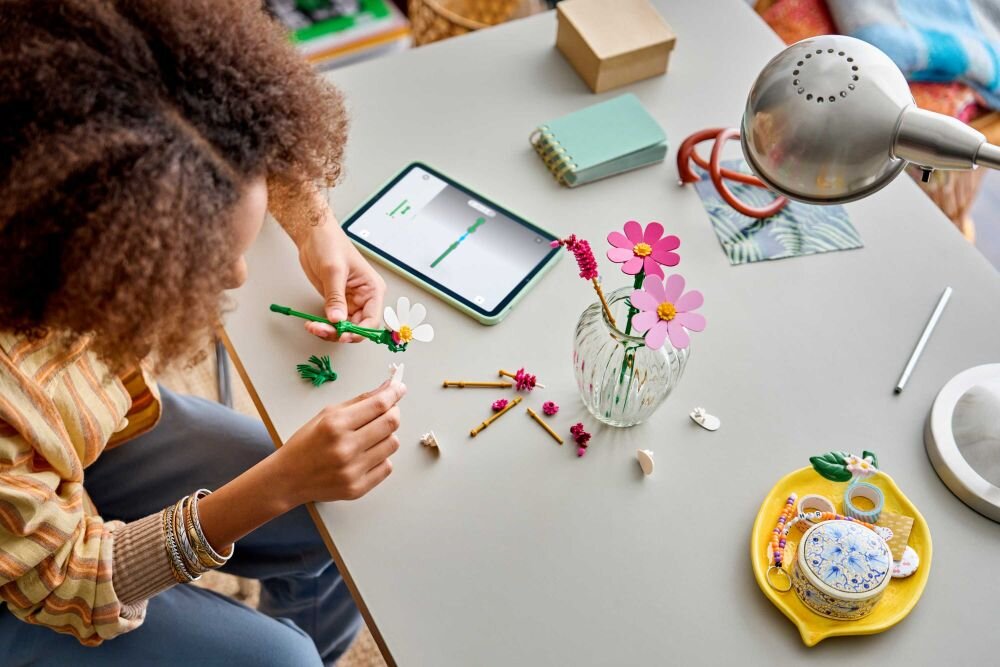 Osoba składa elementy zestawu LEGO Botanicals Kosmosy przy biurku, na którym znajduje się wazon z kwiatami LEGO, tablet, notes i lampka biurkowa. Na biurku widoczne są również drobne akcesoria i ozdoby.
