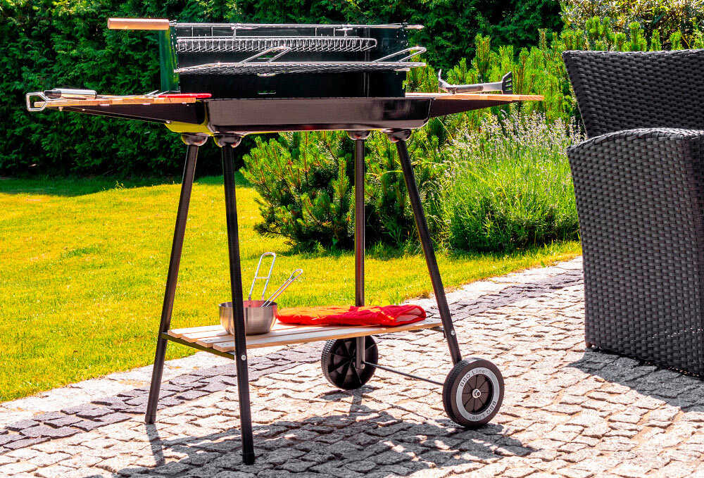 Grill węglowy na kółkach z półkami bocznymi, ustawiony na brukowanej powierzchni w ogrodzie. Na dolnej półce znajdują się akcesoria do grillowania i czerwona tkanina.