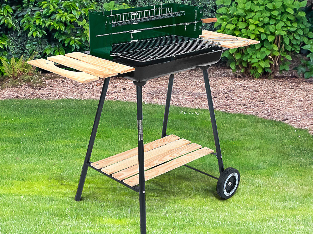Grill węglowy na kółkach z półkami bocznymi i rożnem, ustawiony na trawie w ogrodzie. Grill posiada czarną misę i zieloną osłonę z tyłu.