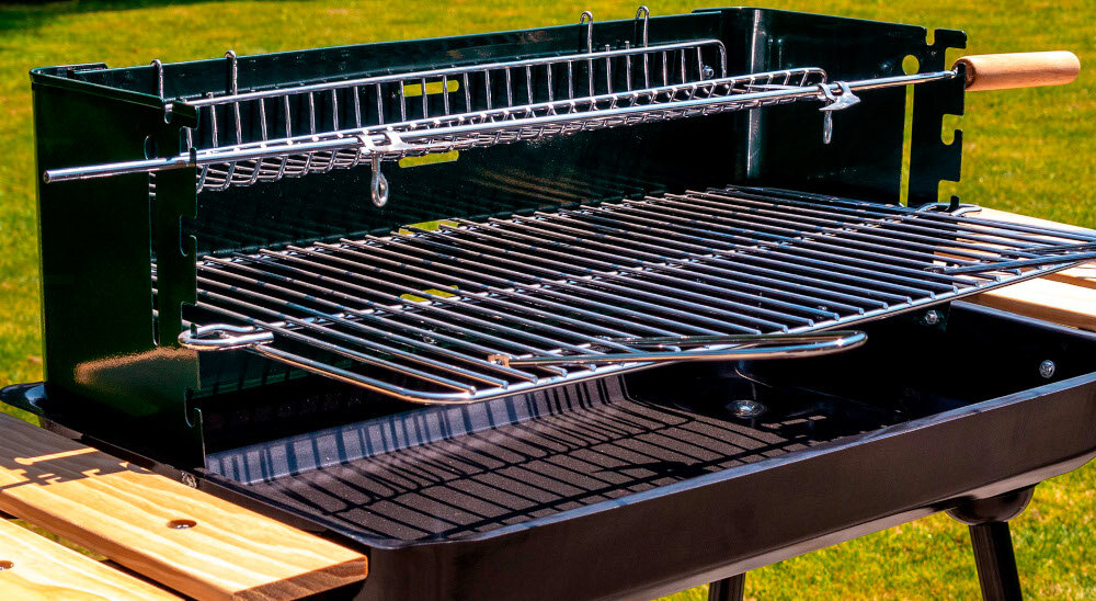 Grill węglowy z metalowym rusztem i rożnem, posiada drewniane półki boczne oraz kółka. Tło stanowi zielona trawa.