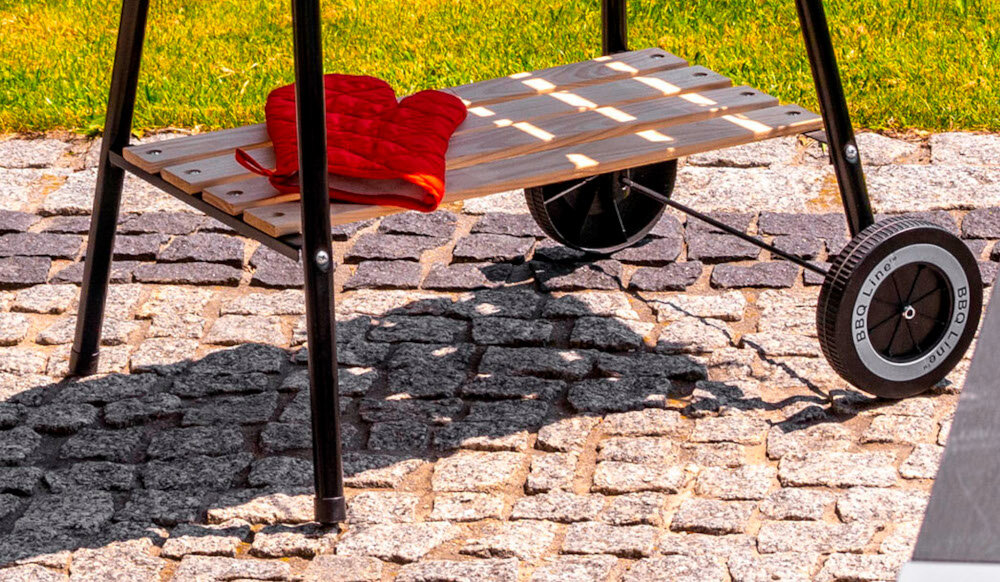 Na obrazie widoczna jest dolna część grilla węglowego z drewnianą półką, na której leży czerwona rękawica. Grill posiada czarne kółka z napisem \'BBQ Line\'.