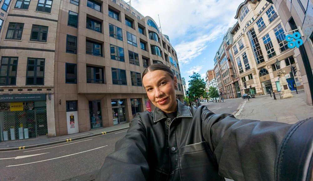 Osoba w ciemnej kurtce robi selfie na tle miejskiej ulicy z nowoczesnymi budynkami. Na jednym z budynków widoczne jest logo \'co-op\'.