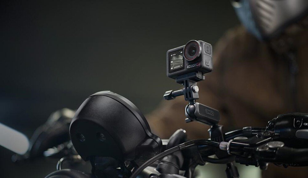Kamera sportowa zamontowana na kierownicy motocykla, w tle widoczna postać w kasku. Kamera ma wyświetlacz z przodu i jest przymocowana za pomocą uchwytu.