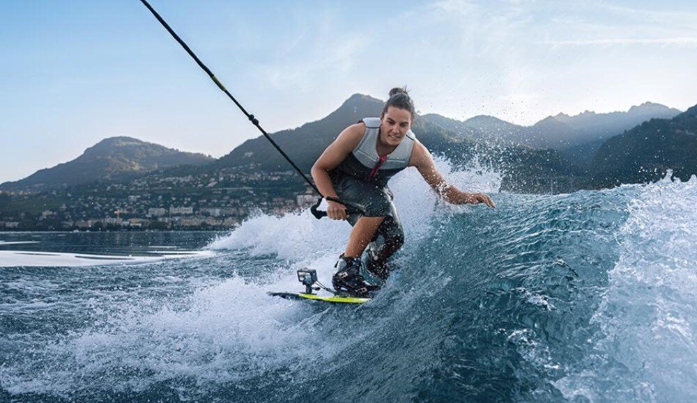 Osoba uprawiająca wakeboarding na jeziorze, otoczona górami w tle. Woda tworzy duże fale, a niebo jest jasne i bezchmurne.
