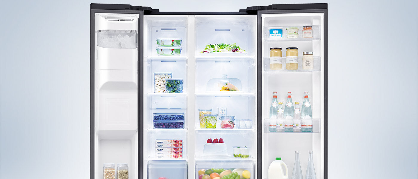 Otwarta lodówka z widocznymi półkami wypełnionymi różnorodnymi produktami spożywczymi, takimi jak owoce, warzywa, napoje i słoiki. W lewej części znajduje się kostkarka do lodu.