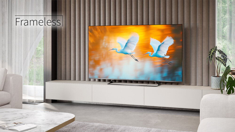 W salonie na szafce stoi telewizor z wyświetlonym obrazem dwóch lecących ptaków na tle pomarańczowego nieba. Na ścianie obok telewizora widoczny jest napis \'Frameless\'.