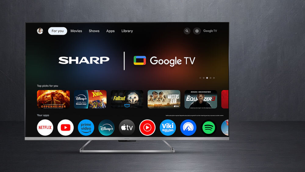 Telewizor wyświetla ekran główny z logo SHARP i Google TV oraz aplikacjami takimi jak Netflix, Prime Video, Disney+, i Spotify. Widoczne są również rekomendacje filmów i seriali.