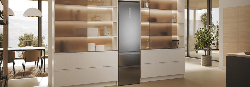 W nowoczesnej kuchni znajduje się lodówka w kolorze inox, otoczona półkami z dekoracjami. W tle widoczny jest stół z krzesłami oraz duże okna z widokiem na zewnątrz.