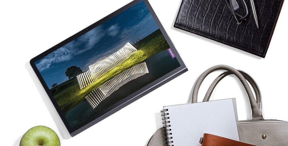 Na obrazie znajduje się tablet z wyświetlaczem pokazującym nowoczesną instalację świetlną na tle krajobrazu. Obok tabletu leży zielone jabłko, notes, długopis, okulary i torba.