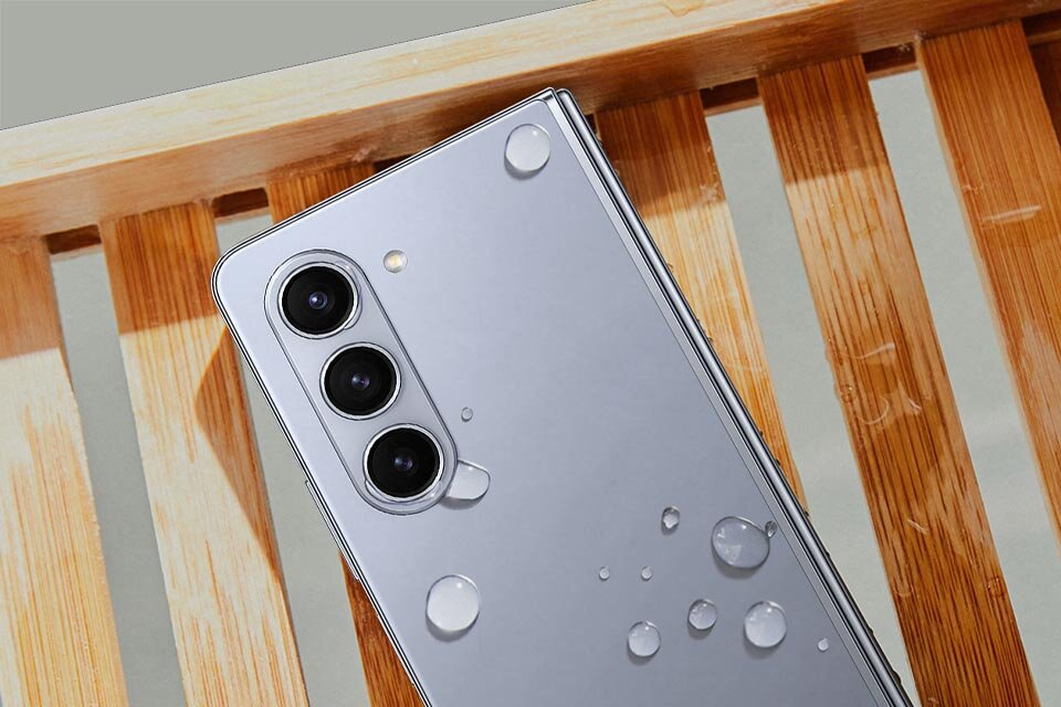Na drewnianej powierzchni leży smartfon z trzema obiektywami aparatu, na którym znajdują się krople wody.