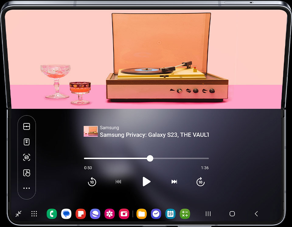 Smartfon z rozłożonym ekranem, na którym wyświetlany jest odtwarzacz multimedialny z utworem \'Samsung Privacy: Galaxy S23, THE VAUL1\'. W tle widoczny jest gramofon oraz dwa kieliszki na różowym tle.