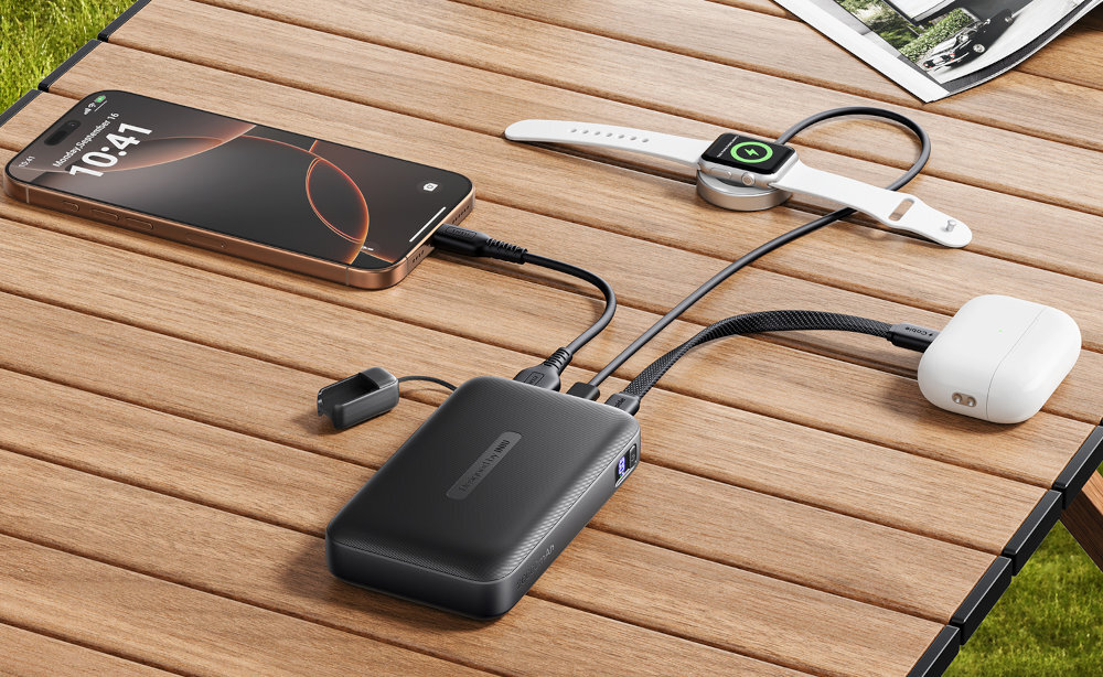 Na drewnianym stole leży czarny powerbank z podłączonymi trzema urządzeniami: smartfonem, smartwatchem i słuchawkami bezprzewodowymi. Powerbank ma widoczne logo producenta i jest w trakcie ładowania urządzeń.