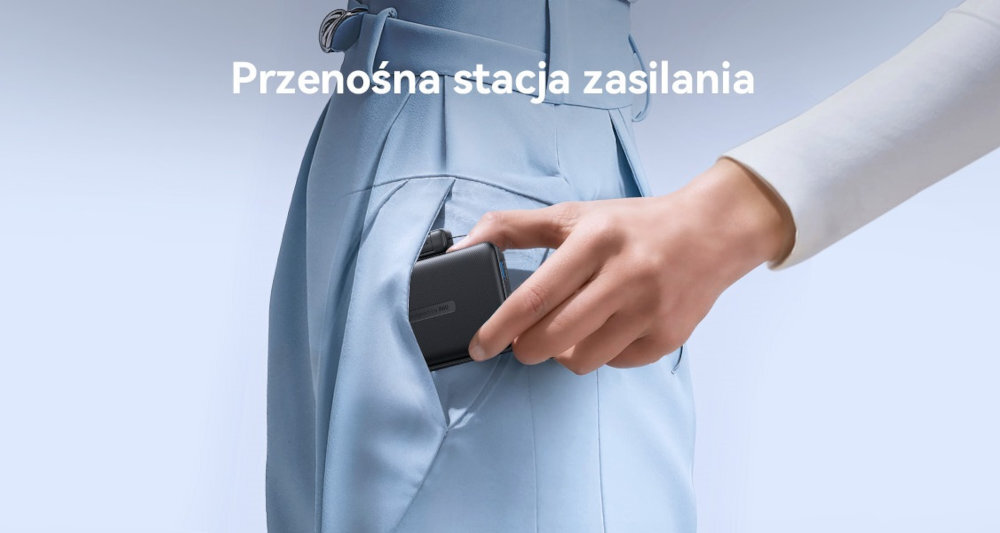 Osoba wkłada czarny powerbank do kieszeni niebieskich spodni. Na obrazie widoczny jest tekst: \'Przenośna stacja zasilania\'.