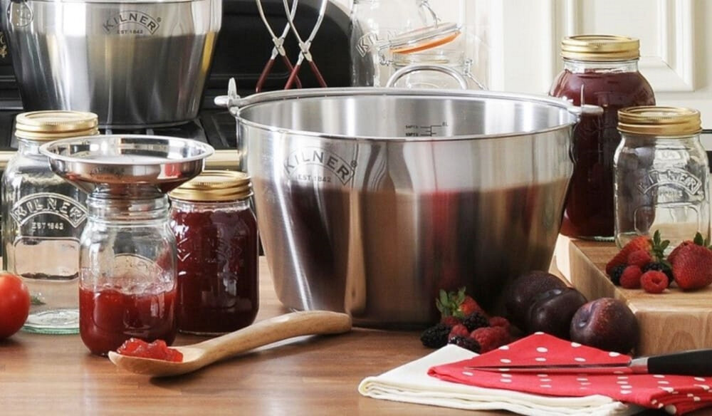 Na drewnianym blacie znajduje się duży metalowy garnek z logo \'KILNER\', otoczony słoikami z przetworami i owocami. Obok leży drewniana łyżka i czerwona ściereczka w białe kropki.