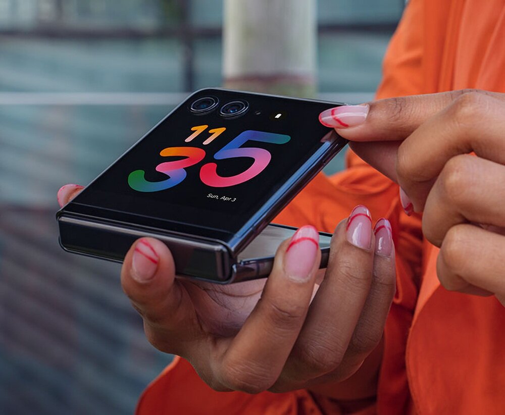 Osoba trzyma składany smartfon z wyświetlaczem pokazującym godzinę 11:35 i datę Sun, Apr 3. Na obudowie widoczne są dwa obiektywy aparatu i małe logo producenta.
