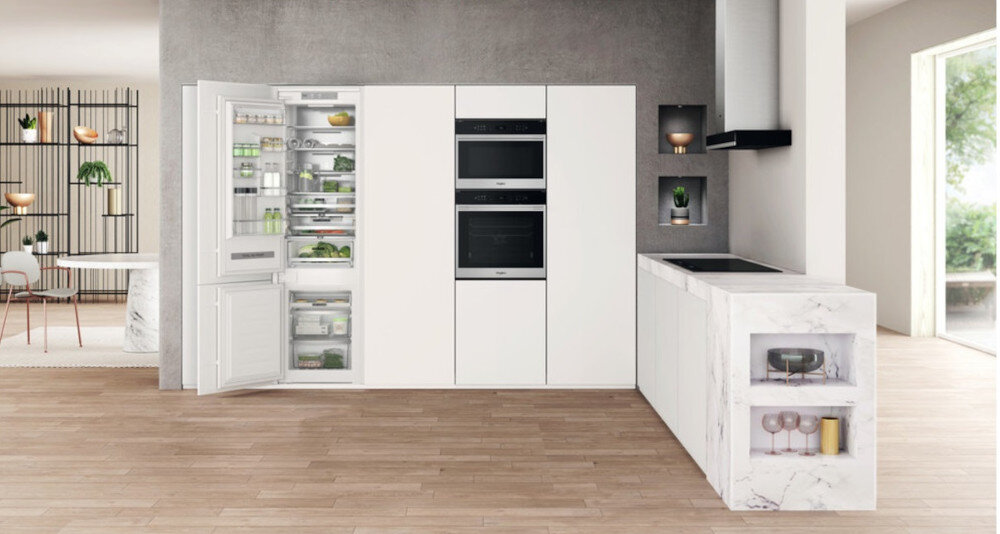 Nowoczesna kuchnia z otwartą lodówką w zabudowie, wypełnioną różnymi produktami spożywczymi. Obok znajdują się dwa piekarniki wbudowane w białą szafkę oraz marmurowy blat z płytą grzewczą.