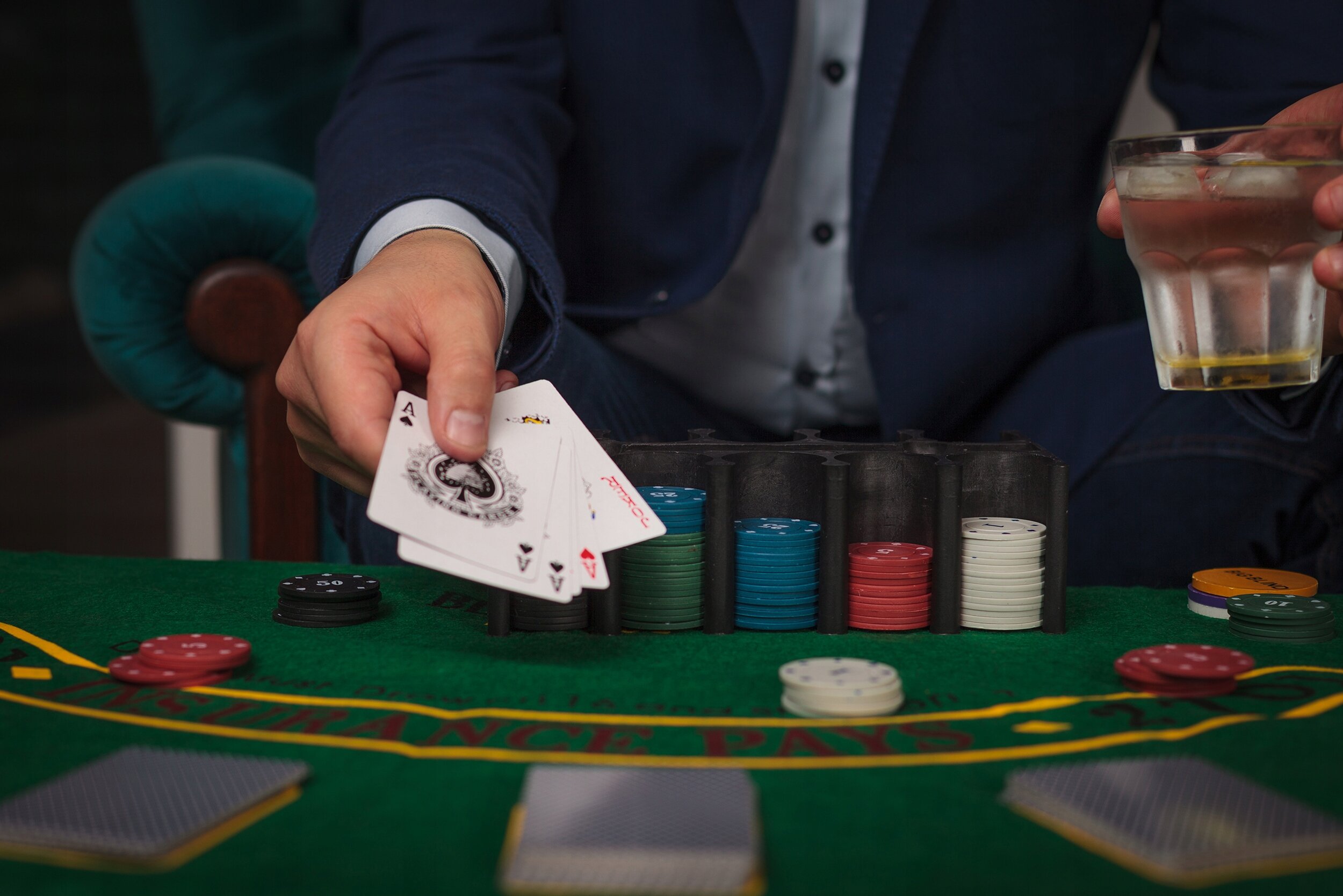 Osoba w garniturze trzyma karty do gry nad zielonym stołem pokerowym z żetonami w różnych kolorach. Obok znajduje się szklanka z napojem.