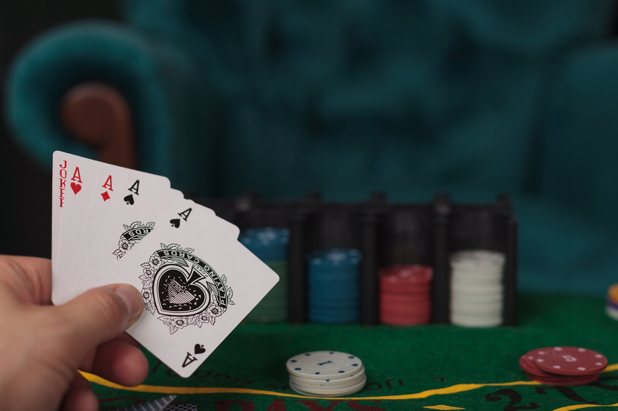 Osoba trzyma w ręku karty do gry: Joker, As kier, As pik, As trefl i As karo. Na stole leżą żetony do pokera w różnych kolorach oraz mata pokerowa.