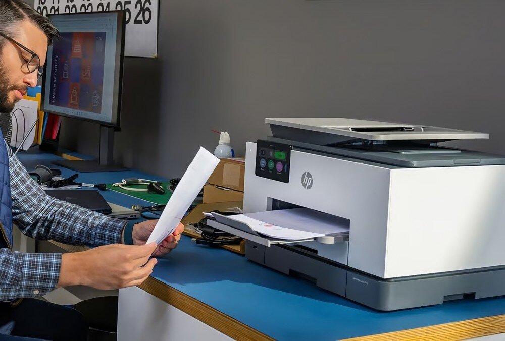 Na biurku znajduje się urządzenie wielofunkcyjne HP z widocznym logo, obok którego siedzi osoba przeglądająca dokumenty. W tle widać monitor z wyświetlonymi ikonami oraz kalendarz na ścianie.