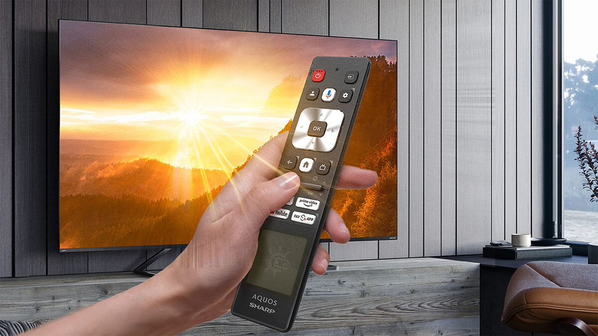 Osoba trzyma pilot z napisem \'AQUOS SHARP\' przed telewizorem wyświetlającym krajobraz z zachodzącym słońcem.